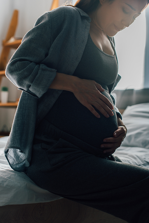 brunette pregnant woman suffering from pain while touching belly in bedroom