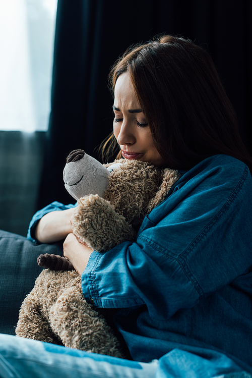 upset brunette woman with closed eyes holding teddy bear in living room
