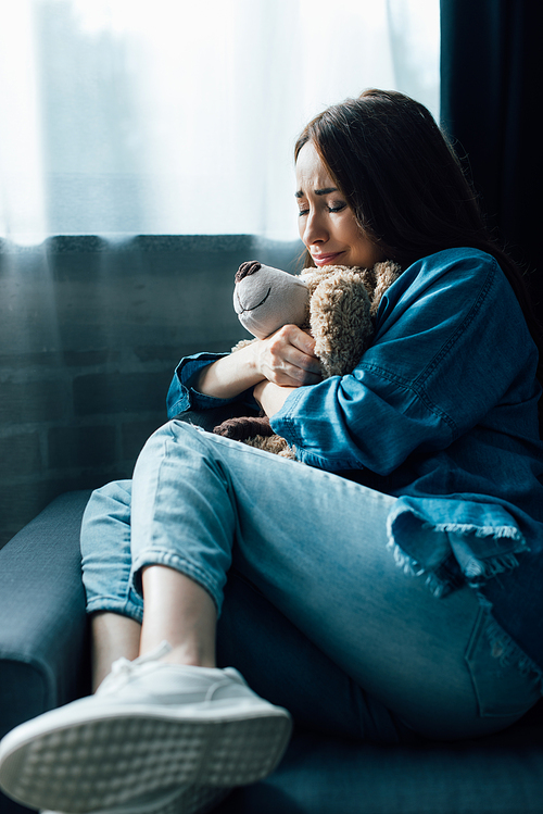 upset brunette woman with closed eyes holding teddy bear while sitting on sofa