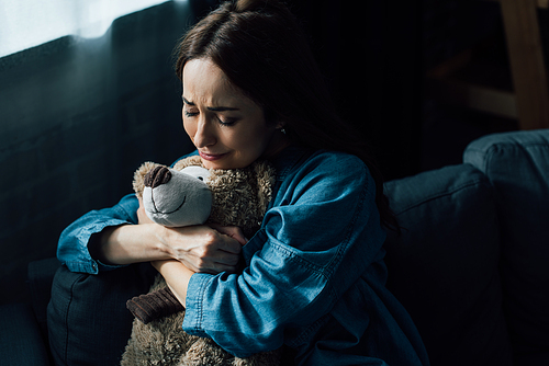 sad brunette woman with closed eyes holding teddy bear in living room