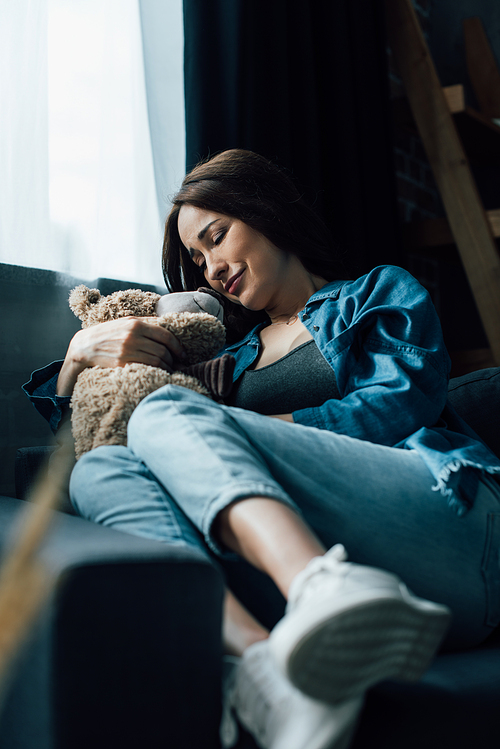 selective focus of upset brunette woman with closed eyes holding teddy bear while sitting on sofa