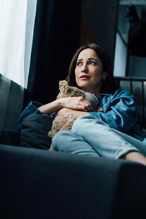 selective focus of depressed brunette woman holding teddy bear while sitting on sofa