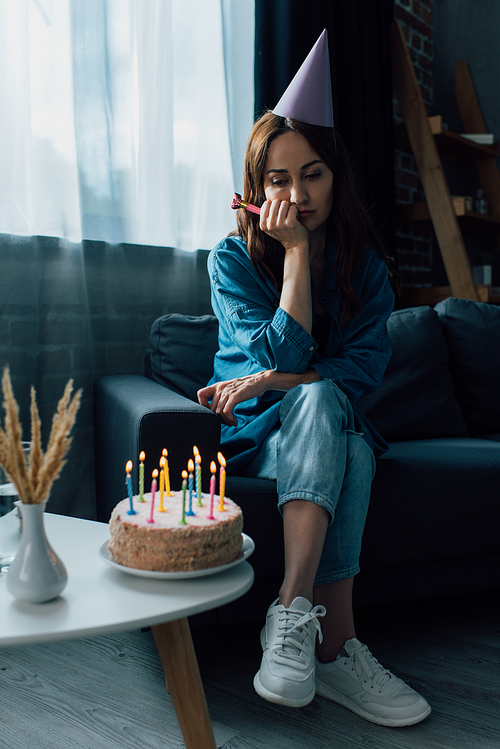 sad woman holding party horn, sitting on sofa and looking at birthday cake on coffee table