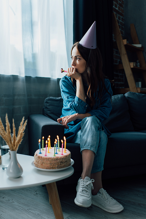 sad woman holding party horn and sitting on sofa near birthday cake on coffee table