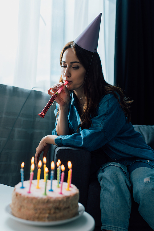 selective focus of sad woman blowing party horn and sitting on sofa near birthday cake on coffee table