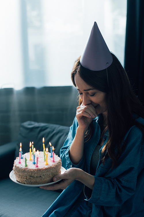 sad woman in party cap crying and holding birthday cake with candles