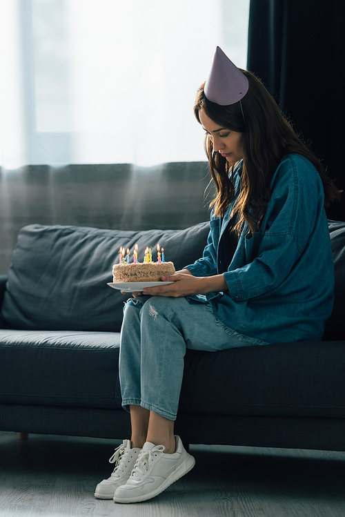 sad woman in party cap sitting on sofa and holding birthday cake with candles