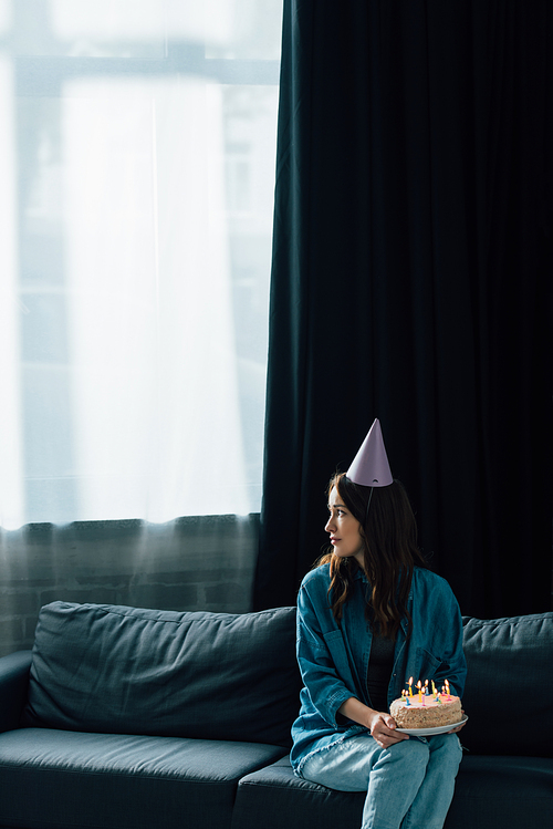 upset woman in party cap sitting on sofa, holding birthday cake with candles and looking away