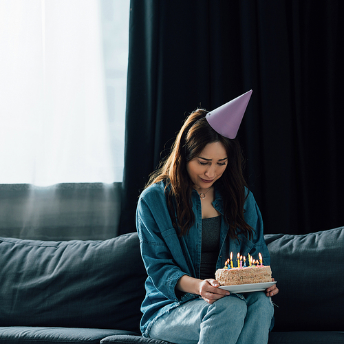 frustrated woman in party cap sitting on sofa and holding birthday cake with candles