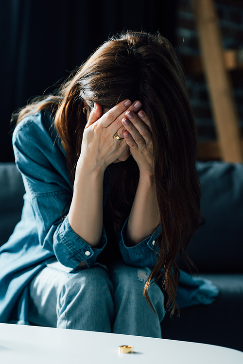 depressed woman covering face while sitting near coffee table with golden ring, divorce concept