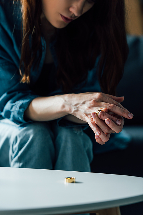 cropped view of brunette woman sitting near coffee table and taking off golden ring, divorce concept