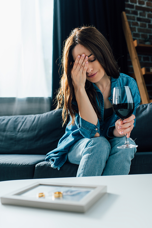 selective focus of depressed woman with glass of wine near golden rings on photo frame, divorce concept