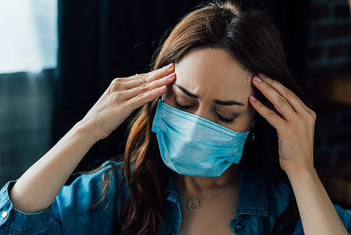 tired brunette woman in medical mask touching head at home