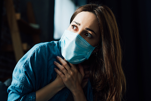 scared brunette woman in blue medical mask touching throat at home