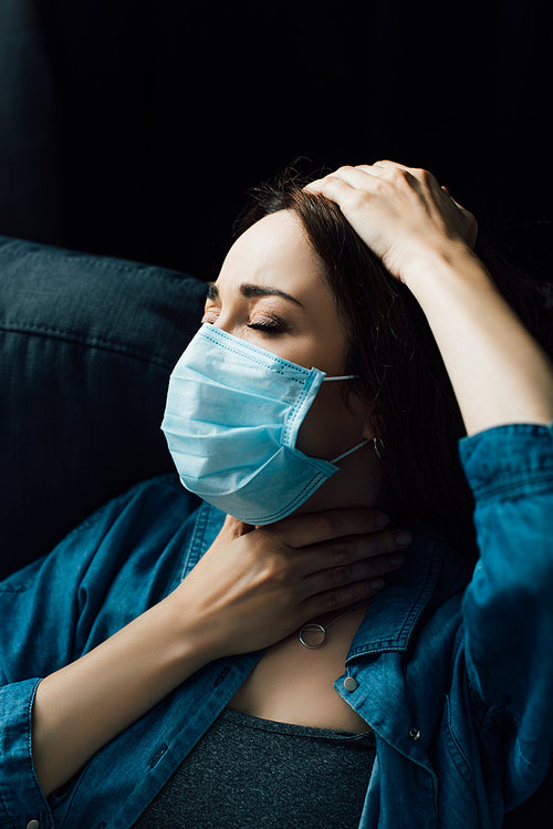 brunette woman with closed eyes in blue medical mask touching throat at home