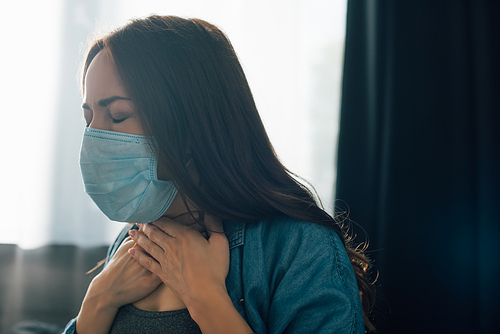 brunette woman with closed eyes in medical mask touching throat at home