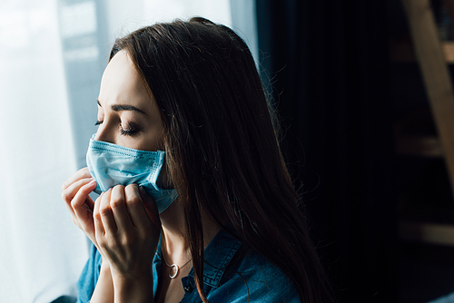 brunette woman with closed eyes touching medical mask at home