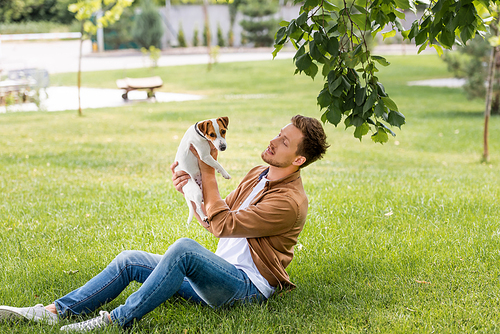 young man in brown shirt and jeans holding jack russell terrier dog while sitting on lawn