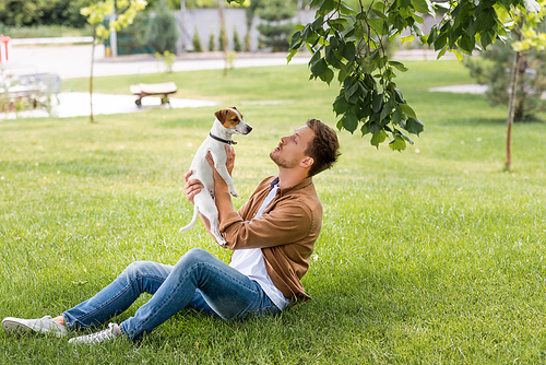 young man in jeans and brown shirt sitting on green grass and holding jack russell terrier dog