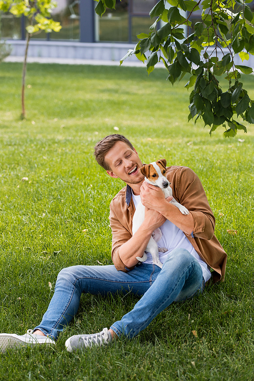 young man in casual clothes sitting on green lawn and cuddling jack russell terrier dog