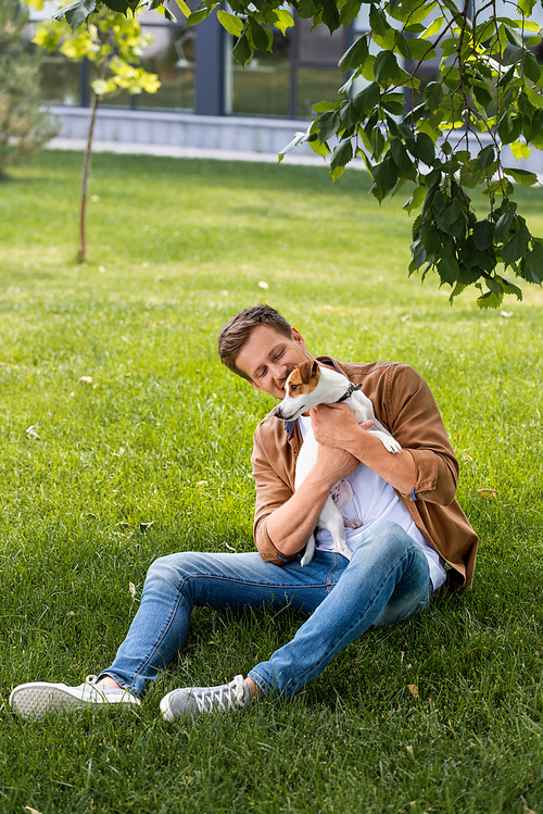 young man in brown shirt and jeans sitting on grass and cuddling jack russell terrier dog