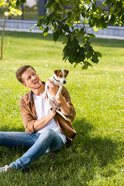 young man in jeans holding jack russell terrier dog while sitting on green grass