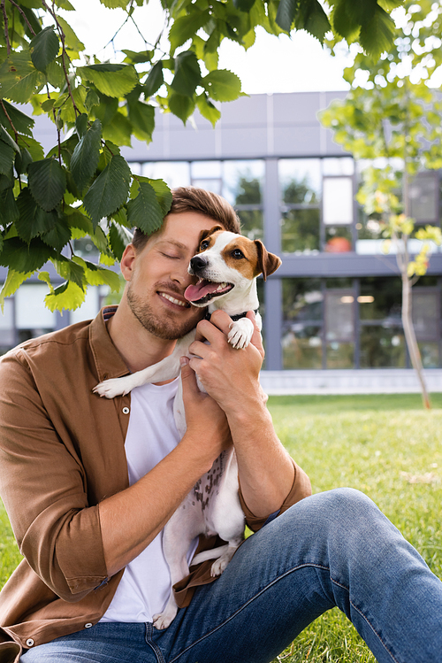 young man in brown shirt and jeans sitting on lawn and cuddling jack russell terrier dog