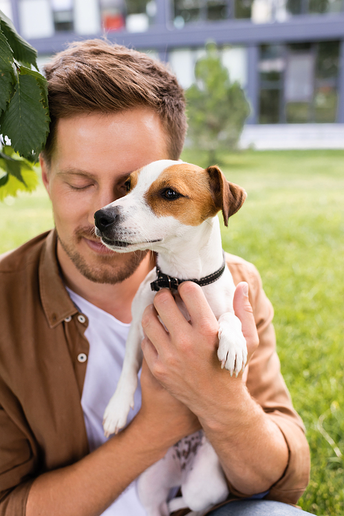 young man with closed eyes holding jack russell terrier dog with brown spots on head