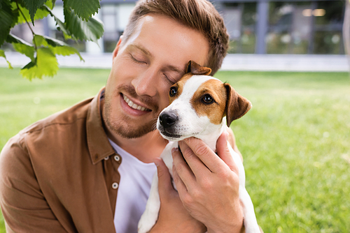 young man with closed eyes holding white jack russell terrier dog with brown spots on head