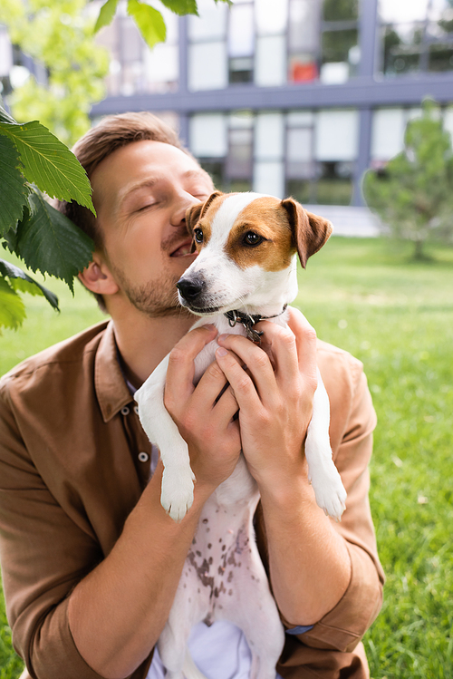 young man with closed eyes holding white jack russell terrier dog with brown spots on head