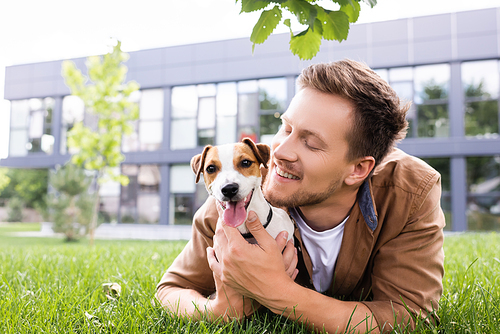 young man cuddling jack russell terrier dog while lying on green lawn
