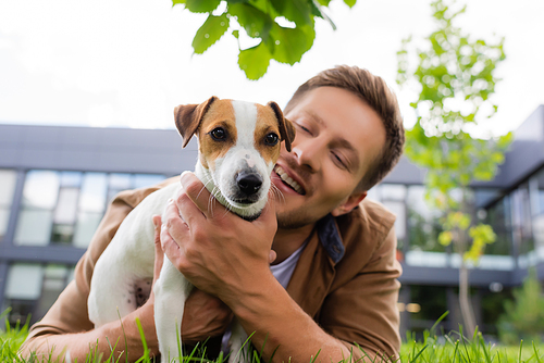 selective focus of young man cuddling jack russell terrier dog while lying on green grass