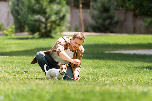Selective focus of young man kneeling near jack russell terrier on leash in park