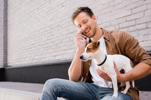 Young man talking on smartphone and hugging jack russell terrier on sidewalk near building