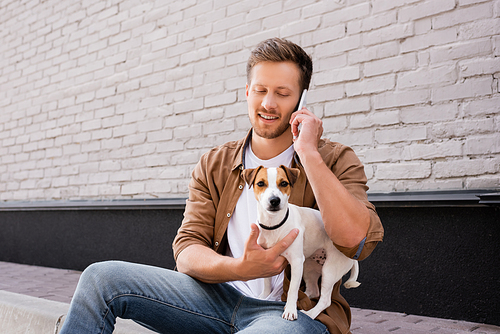 Man hugging jack russell terrier and talking on smartphone on sidewalk near facade of building
