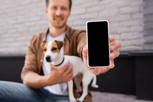 Selective focus of man showing smartphone with blank screen and embracing jack russell terrier on urban street