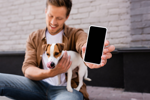 Selective focus of man showing smartphone with blank screen while holding jack russell terrier on urban street