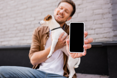 Selective focus of young man holding smartphone with blank screen and jack russell terrier on urban street