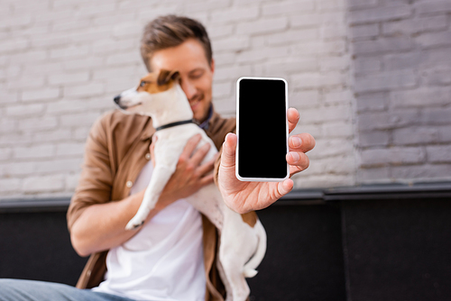 Selective focus of man holding smartphone with blank screen and hugging jack russell terrier on urban street