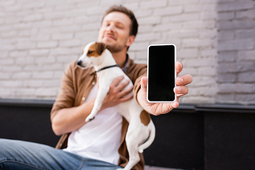 Selective focus of young man holding cellphone with blank screen and jack russell terrier on urban street