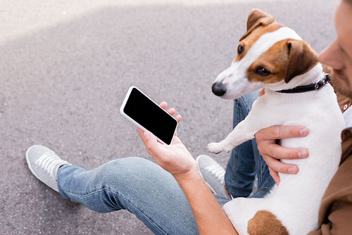 Selective focus of man holding smartphone with blank screen and hugging jack russell terrier outdoors