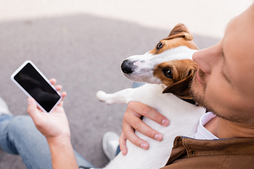 Selective focus of man embracing jack russell terrier and holding smartphone with blank screen outdoors