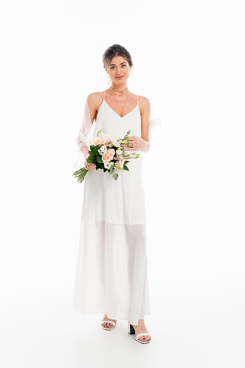 full length view of smiling woman in wedding dress holding bouquet on white