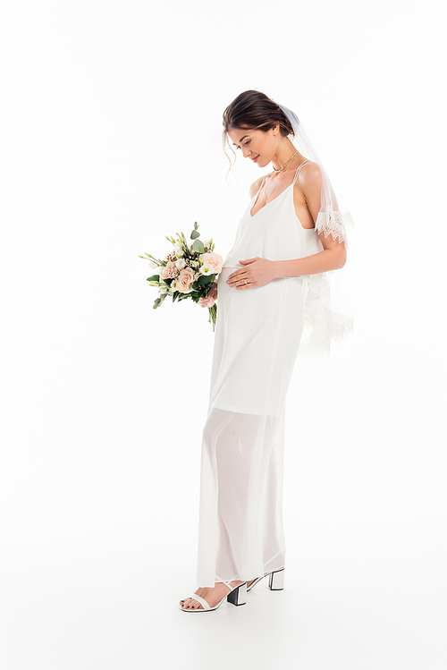 full length view of pregnant bride touching belly while holding bouquet on white