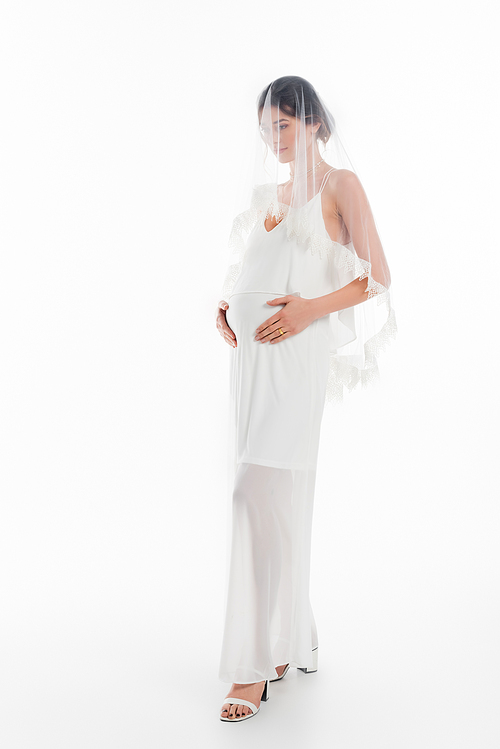 full length view of pregnant bride in veil touching tummy on white