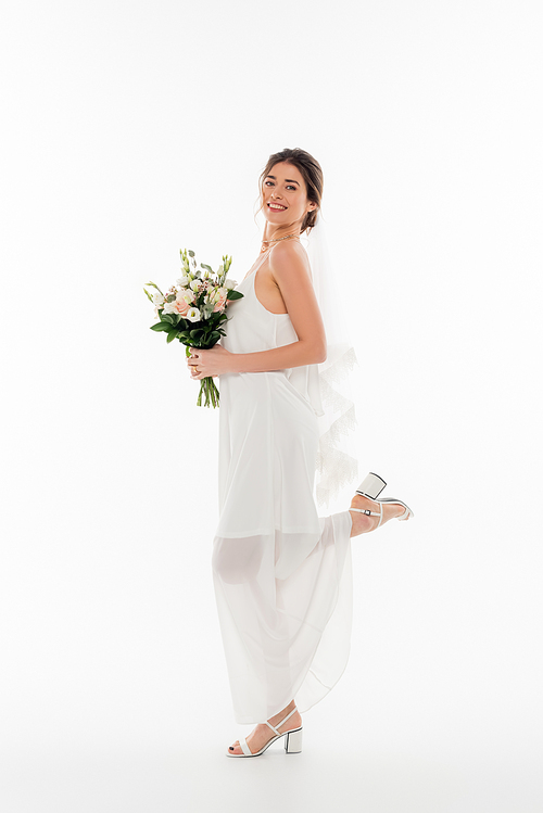 full length view of joyful woman in wedding dress posing with wedding bouquet on white