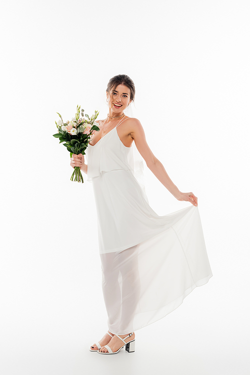 full length view of happy fiancee with wedding bouquet smiling at camera on white