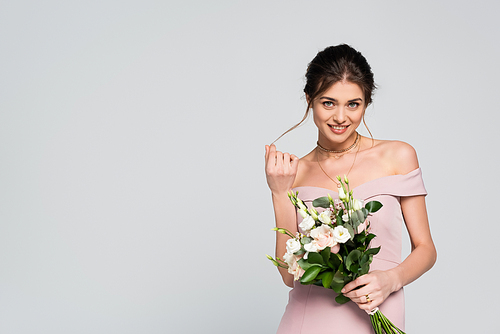 flirty woman touching hair and smiling at camera while holding wedding bouquet isolated on grey