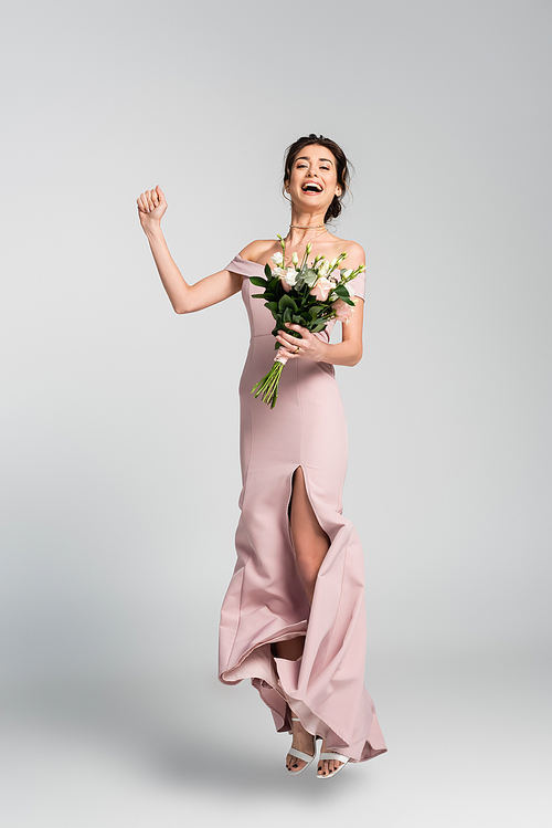 full length view of excited fiancee holding wedding bouquet while jumping on grey