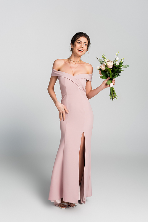 full length view of excited woman in long wedding dress holding bouquet and looking away on grey
